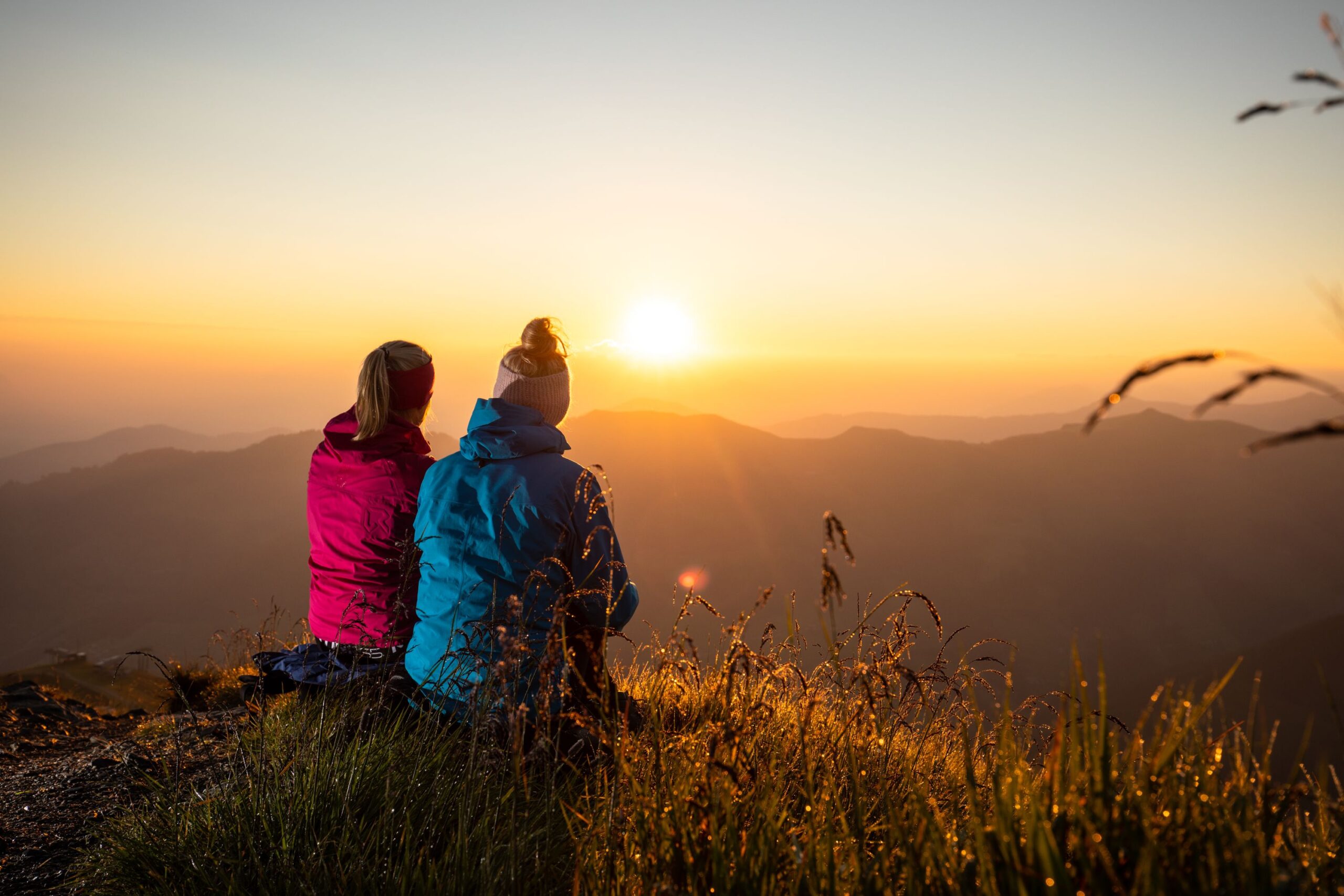 zwei Damen im Gras sitzend am Berg beobachten einen orange-gelben Sonnenaufgang hinter den Bergen