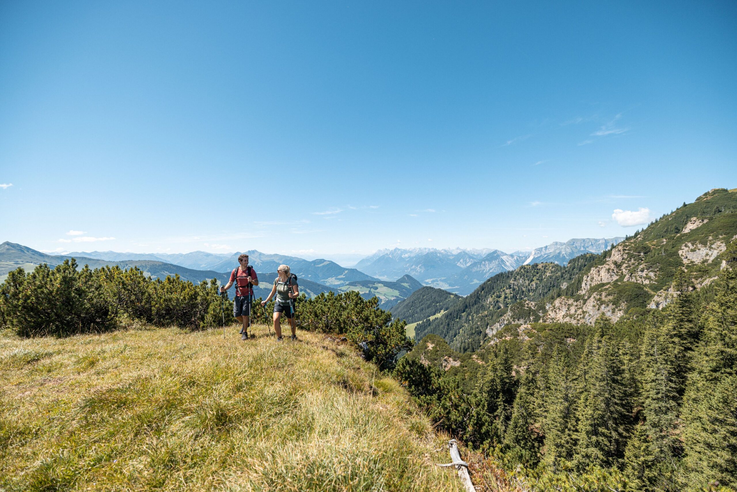 Junges Pärchen wandert mit Wanderstöcken im Gras im Hintergrund Latschen, Berge und viel Grün
