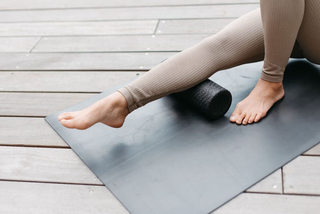 Beine einer Person mit grauen Leggins auf einer Yogamatte und einer Faszienrolle unter der Wade